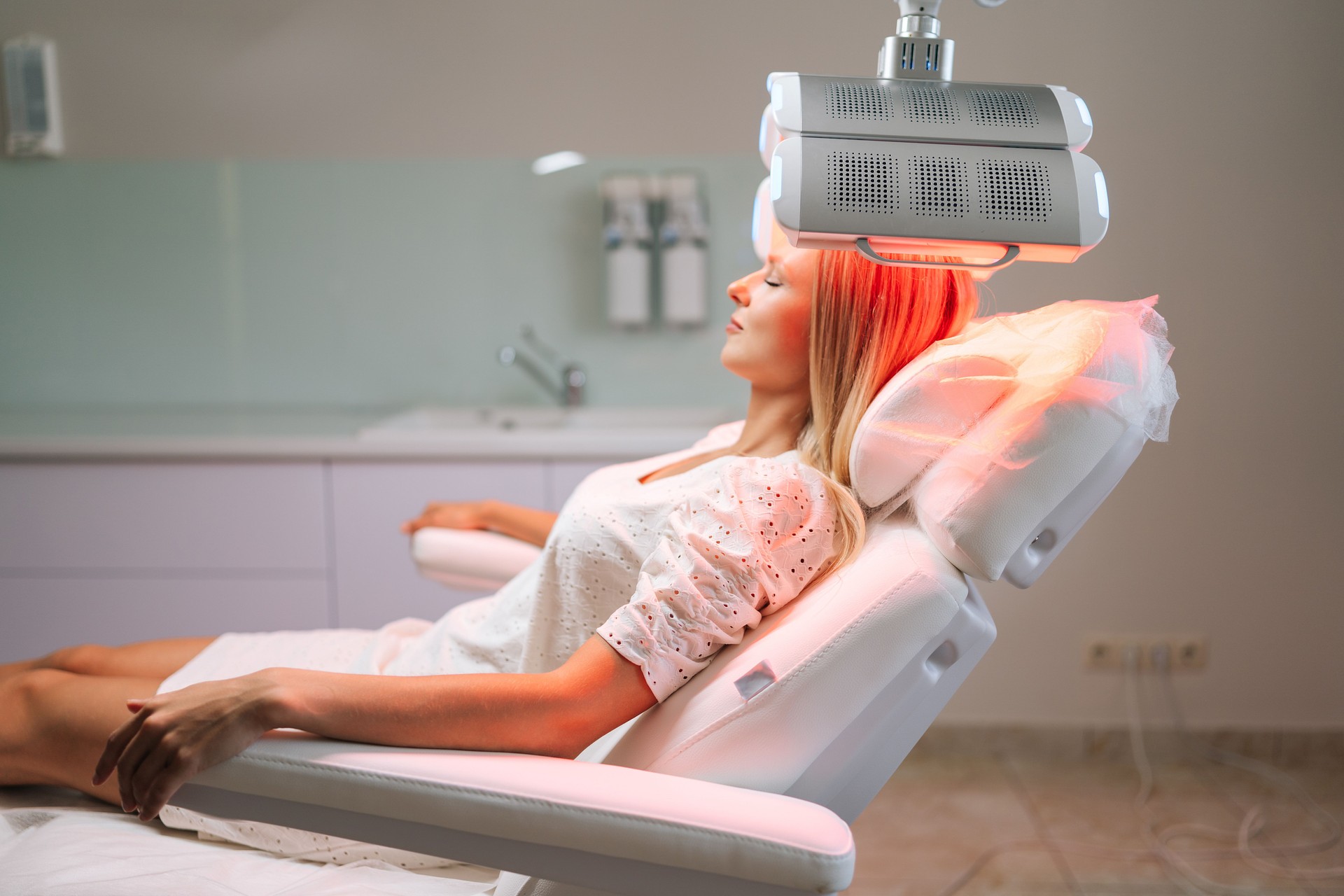 Relaxed blonde female with closed eyes lying on procedure chair having LED red light hair photodynamic therapy during hair treatment at medical clinic.